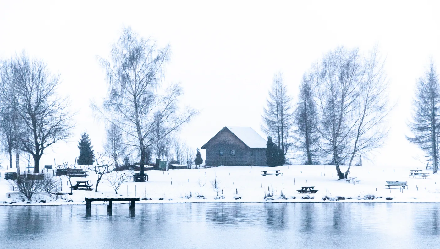 A wooden hut among bare trees on the snowy shore of the Wolfgangsee, with a small jetty on the water
